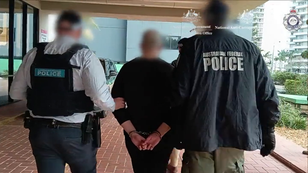A man is led away in handcuffs by two uniformed police officers, with their faces blurred from behind.